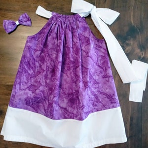 May include: A purple and white sleeveless dress with a white bow tie. The dress has a gathered neckline and a white bow at the back.