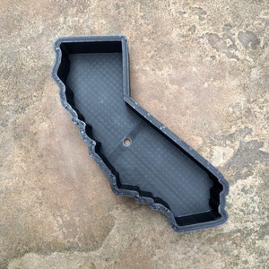 May include: A black, 3D-printed tray shaped like the state of California. The tray features a textured, grid-like base, raised walls with a layered finish, and a single circular hole located near the center of the base, providing a distinct outline.