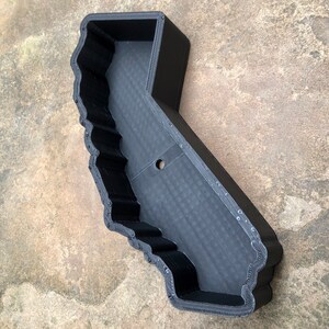 May include: A black, 3D-printed tray shaped like the state of California. The object features a hollow interior with a flat base and raised, textured walls that follow the geographic outline of the state, including a single circular hole in the center.