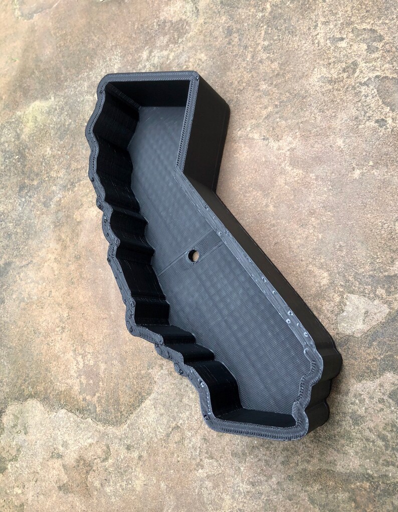 May include: A black, 3D-printed tray shaped like the state of California. The object features a hollow interior with a flat base and raised, textured walls that follow the geographic outline of the state, including a single circular hole in the center.