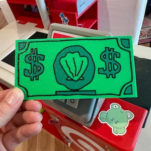 Puede incluir: Un billete de juguete rectangular de color verde con un contorno negro brillante, dos signos de dólar en los laterales y un emblema circular central que representa una concha. El dinero de juguete tiene una superficie texturizada ante una caja.