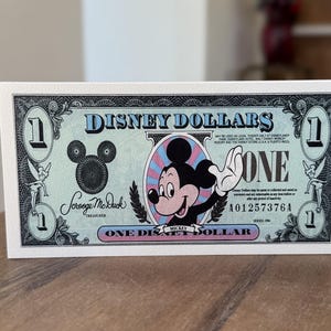 May include: A rectangular Disney Dollars one-dollar bill print featuring a light green background, a central illustration of Mickey Mouse, and the text Disney Dollars, One, and Scrooge McDuck Treasurer. The design includes Tinker Bell figures.