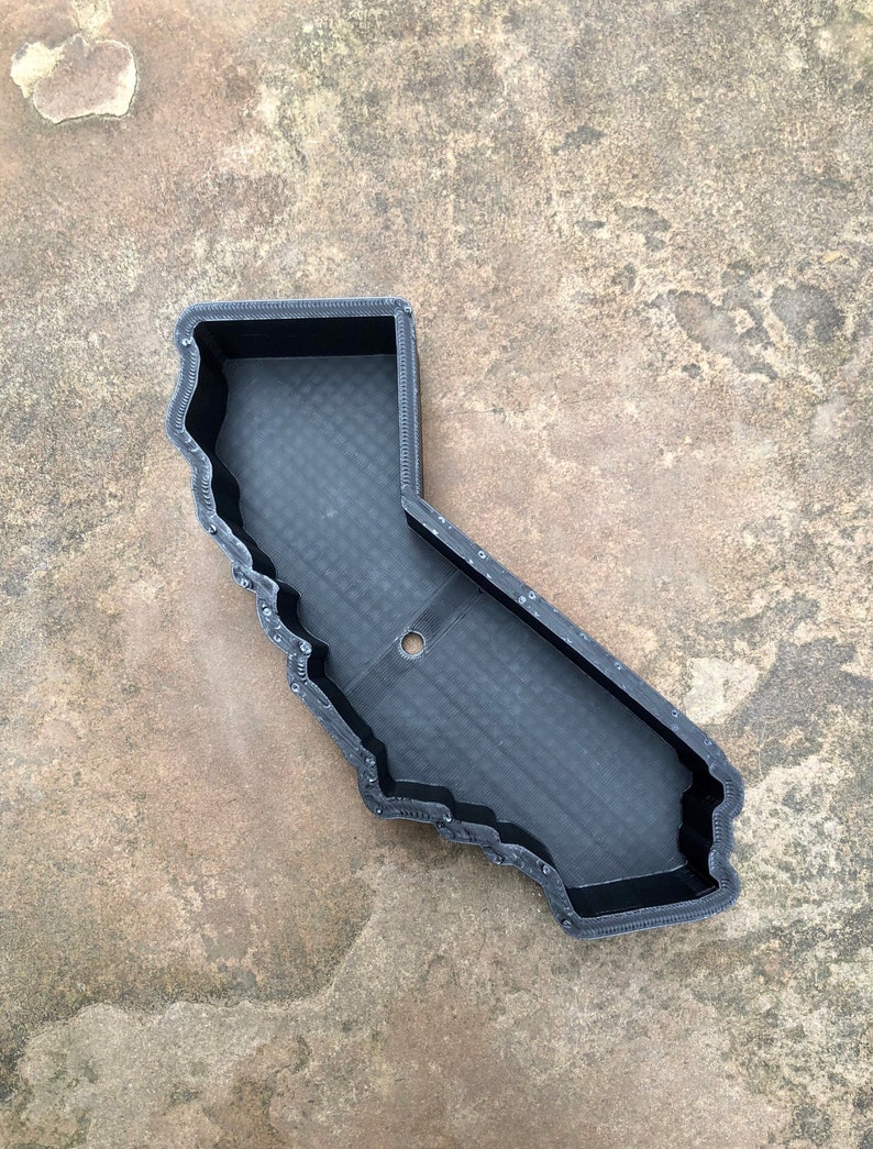 May include: A black, 3D-printed tray shaped like the state of California. The tray features a textured, grid-like base, raised walls with a layered finish, and a single circular hole located near the center of the base, providing a distinct outline.