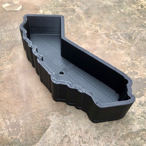 May include: A black, 3D-printed tray shaped like the outline of the state of California. The container features high, vertical walls with a textured, layered finish and a single circular drainage hole located on the flat base of the interior floor.