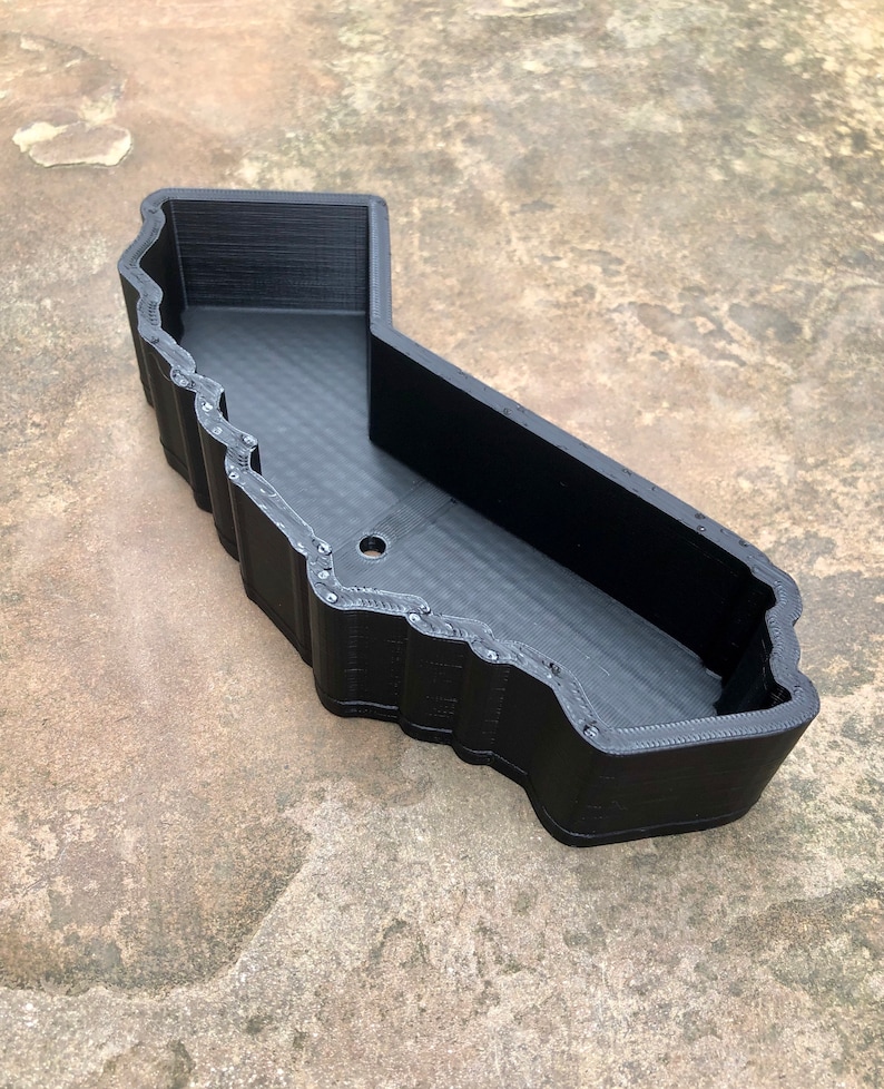 May include: A black, 3D-printed tray shaped like the outline of the state of California. The container features high, vertical walls with a textured, layered finish and a single circular drainage hole located on the flat base of the interior floor.