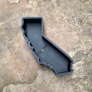 May include: A black, 3D-printed tray shaped like the state of California. The object features a raised outer wall and a flat, textured base with a single circular hole located in the center. The surface of the tray displays visible layer lines.
