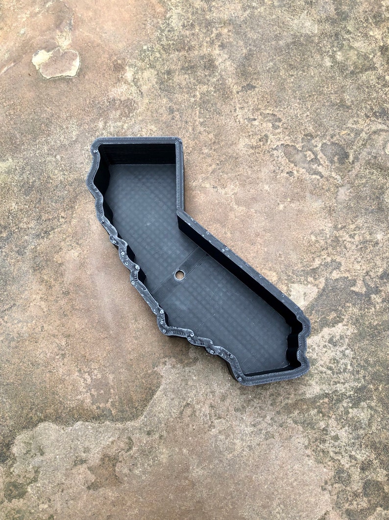 May include: A black, 3D-printed tray shaped like the state of California. The object features a raised outer wall and a flat, textured base with a single circular hole located in the center. The surface of the tray displays visible layer lines.