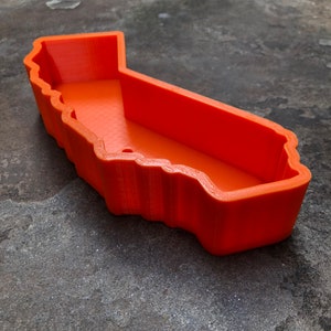 May include: A bright orange, 3D-printed plastic tray shaped like the outline of the state of California. The container features high, vertical walls with a textured finish and a flat, open interior base, creating a hollow, map-shaped vessel.