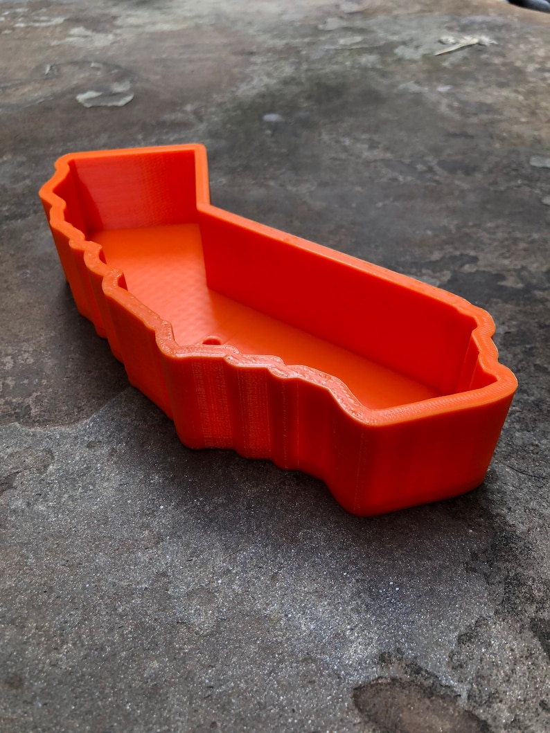 May include: A bright orange, 3D-printed plastic tray shaped like the outline of the state of California. The container features high, vertical walls with a textured finish and a flat, open interior base, creating a hollow, map-shaped vessel.