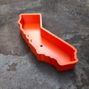 May include: A bright orange, hollow container shaped like the state of California. The object features a distinct, raised border following the geographic outline of the state, with a smooth interior base and a single small circular hole near the center.