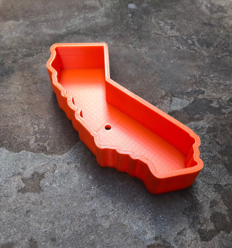 May include: A bright orange, hollow container shaped like the state of California. The object features a distinct, raised border following the geographic outline of the state, with a smooth interior base and a single small circular hole near the center.