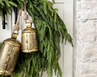 Op de afbeelding: Een feestelijke kerstkrans met kunstmatige dennentakken en twee goudkleurige metalen bellen. De bellen zijn opgehangen aan natuurlijke touwen en hebben een textuur. De krans wordt getoond tegen een witte achtergrond en een stenen muur.