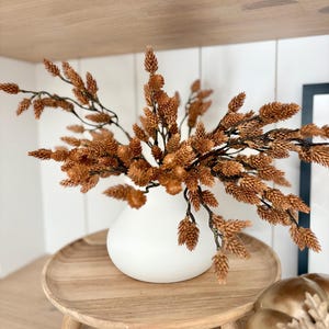 May include: A white ceramic vase containing a cluster of dried brown pinecone-like stems with dark, thin branches. The vase has a rounded, bulbous shape and sits on a circular wooden pedestal tray with three short legs, creating a natural display.