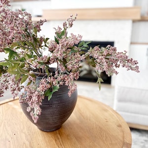 May include: A dark brown ceramic vase filled with pink and green floral branches. The vase sits on a light brown wooden table. The background includes a white fireplace and a white chair.