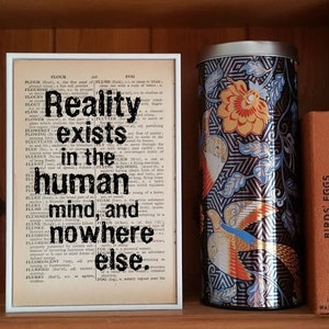 Pode incluir: Uma impressão emoldurada branca com um fundo de página de dicionário em preto e branco. O texto "Reality exists in the human mind, and nowhere else." está impresso em preto na página. A impressão está colocada em uma prateleira de madeira com um copo de metal e dois livros.