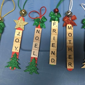 May include: Five wooden ornaments decorated with green glitter and red bows. Each ornament has a word spelled out using letter tiles. The words are "Sister", "Joy", "Noel", "Friend", "Mother", and "Dad".