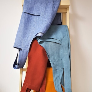 Może przedstawiać: Trzy lniane fartuchy wiszące na drewnianej drabinie. Fartuchy są w odcieniach niebieskiego, pomarańczowego i czerwonego.