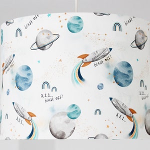 Może przedstawiać: Biały klosz lampy z wzorem o tematyce kosmicznej. Wzór przedstawia rakiety, planety, gwiazdy i tęcze. Tekst "3,2,1... blast off!" powtarza się w całym wzorze.