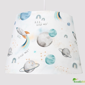 Op de afbeelding: Een witte lampenkap met een ruimtethema. Het heeft een raket, planeten, sterren en regenbogen. De tekst "3, 2, 1... blast off!" staat op de lampenkap.