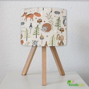 Puede incluir: Una lámpara de mesa de madera con una pantalla beige con estampado de animales del bosque. La pantalla tiene un patrón de zorros, búhos, erizos, setas y árboles. La lámpara tiene una base de trípode.