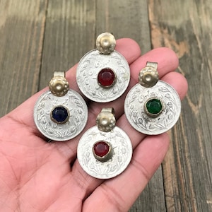 Puede incluir: Cuatro colgantes de moneda de plata con piedras de vidrio de diferentes colores en el centro. Las piedras son roja, azul, verde y roja. Los colgantes est&aacute;n en una mano.