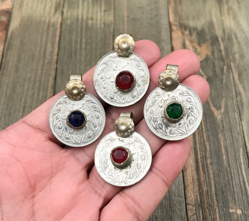 Puede incluir: Cuatro colgantes de moneda de plata con piedras de vidrio de diferentes colores en el centro. Las piedras son roja, azul, verde y roja. Los colgantes est&aacute;n en una mano.