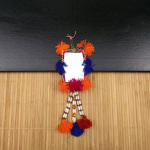 Puede incluir: Un pequeño espejo enmarcado con fondo blanco y un borla de colores colgando de la parte inferior. El marco está decorado con pompones y cuentas de colores.