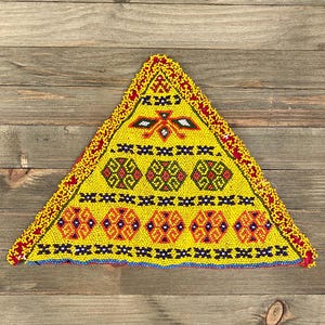 Puede incluir: Un trozo de tela triangular con un patrón geométrico hecho de cuentas amarillas, rojas, azules, verdes y negras. La tela está flecos en los bordes.