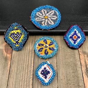 May include: Five round and square beaded ornaments with blue, yellow, white, and red beads. The ornaments are on a wooden surface.