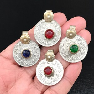 Puede incluir: Cuatro colgantes de moneda de plata con piedras de vidrio de diferentes colores en el centro. Las piedras son roja, verde, azul y roja. Los colgantes est&aacute;n en una mano.