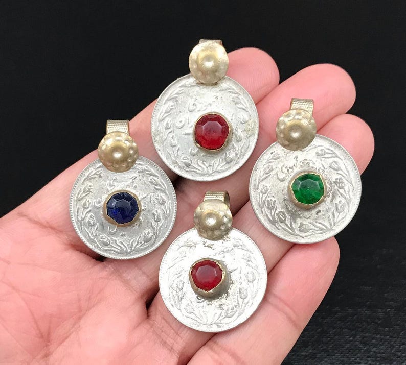 Puede incluir: Cuatro colgantes de moneda de plata con piedras de vidrio de diferentes colores en el centro. Las piedras son roja, verde, azul y roja. Los colgantes est&aacute;n en una mano.