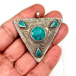 Puede incluir: Un colgante triangular de plata con diseños intrincados y piedras turquesa. El colgante tiene un bucle en la parte superior para sujetarlo a una cadena.