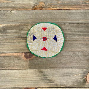May include: A round, white, embroidered fabric with a green border. The embroidery features a geometric pattern in yellow, red, and blue.