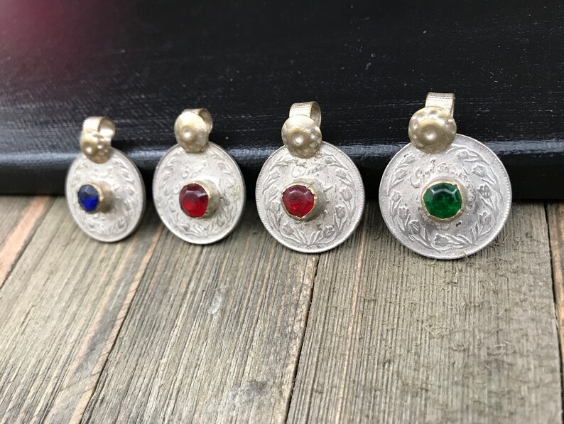 Puede incluir: Cinco colgantes de monedas de plata con piedras de vidrio de diferentes colores en el centro. Las monedas tienen escritura &aacute;rabe alrededor del borde.