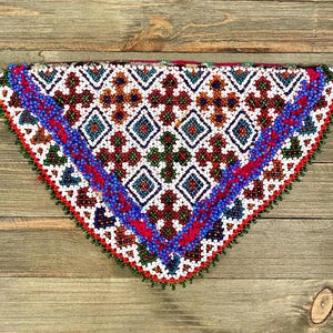 Puede incluir: Una pieza triangular de tela con un patrón geométrico de cuentas blancas, rojas, azules y verdes. La tela está bordeada con un ribete de cuentas rojas y azules.