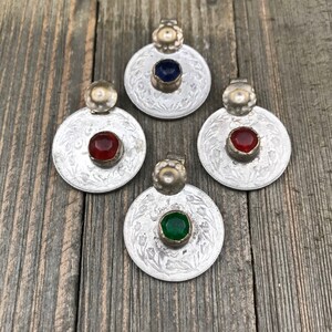 Puede incluir: Cuatro colgantes de moneda de plata con piedras de vidrio de diferentes colores. Las piedras son azules, rojas, verdes y rojas. Los colgantes est&aacute;n sobre una superficie de madera.
