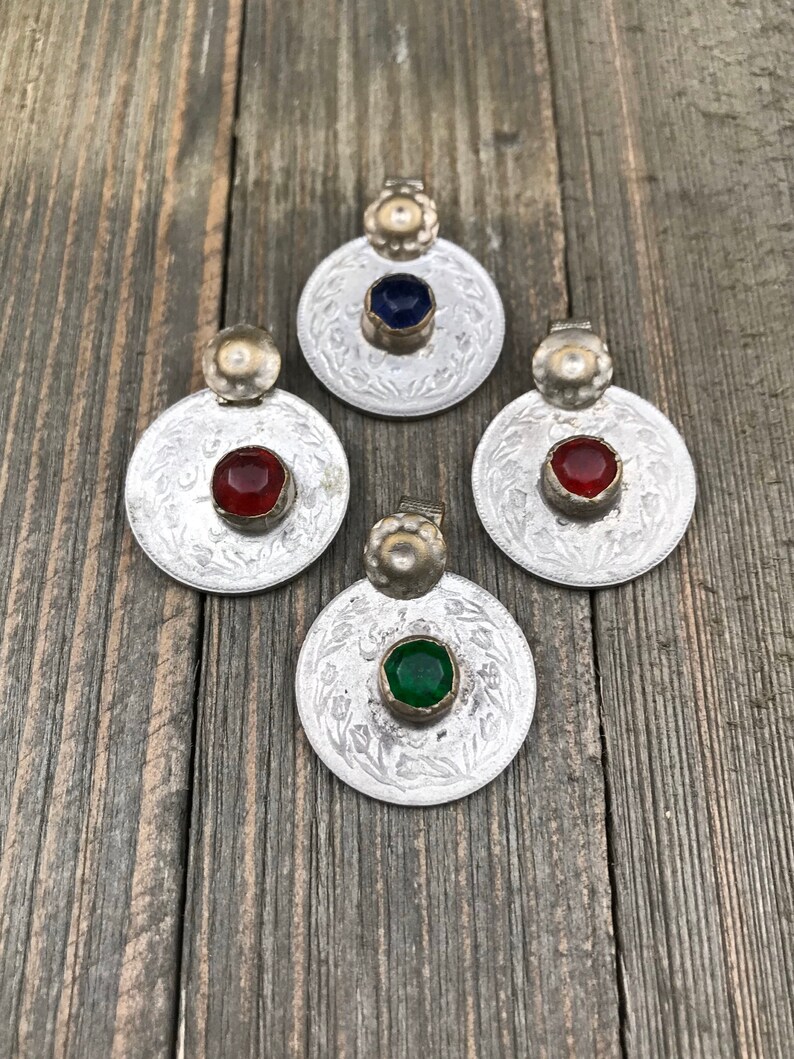 Puede incluir: Cuatro colgantes de moneda de plata con piedras de vidrio de diferentes colores. Las piedras son azules, rojas, verdes y rojas. Los colgantes est&aacute;n sobre una superficie de madera.