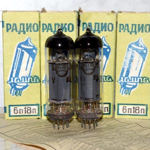 Op de afbeelding: Twee vintage vacuümbuizen, 6N18n, in hun originele dozen. De dozen zijn geel met blauwe en witte tekst die "RADIO ЛАМПА 6N18n" luidt.