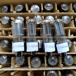 May include: A collection of clear glass vacuum tubes arranged in a cardboard grid. Four tubes in the front row feature white paper labels with the handwritten text Ia=132mA or J=132mA attached to their black bases, revealing internal metal components.