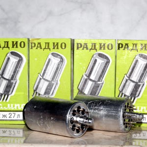 Peut inclure: Quatre boîtes de tubes à vide vintage étiquetées "RADIO" en écriture cyrillique. Les boîtes sont vertes avec des lettres blanches et présentent des illustrations des tubes. Deux tubes à vide argentés sont visibles au premier plan.