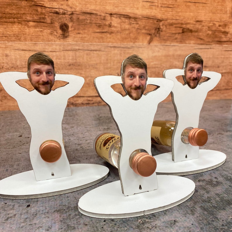 May include: Three white wooden bottle holders shaped like a person with a face cutout. Each holder has a small bottle of liquor inserted into the cutout. The bottles are clear with a gold cap.