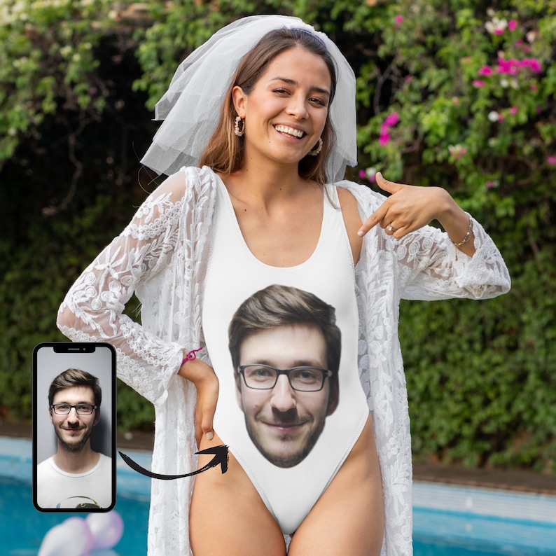 May include: A white swimsuit with a man's face printed on the front. The swimsuit has a lace overlay and a white veil on the head of the person wearing it.