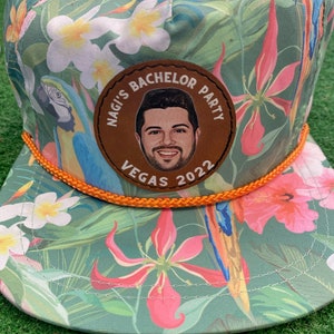 May include: A green, tropical print baseball cap with an orange rope trim. A brown leather patch with the text "Nagi's Bachelor Party Vegas 2022" and a photo of a man is sewn onto the front of the cap.