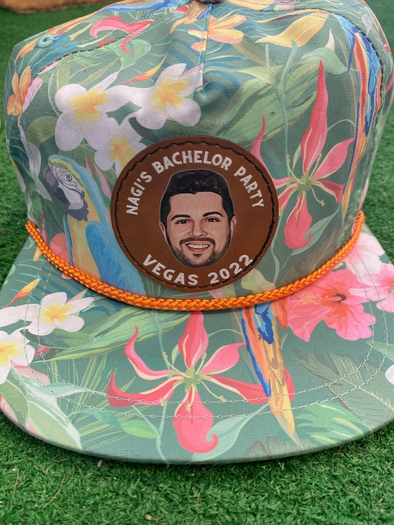 May include: A green, tropical print baseball cap with an orange rope trim. A brown leather patch with the text "Nagi's Bachelor Party Vegas 2022" and a photo of a man is sewn onto the front of the cap.