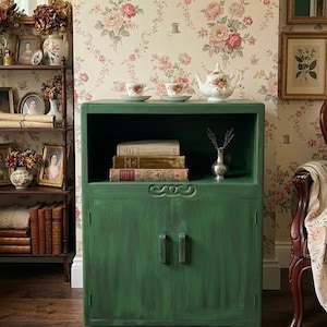 May include: A dark green cabinet with a shelf and two doors, holding books and a small vase. Two teacups and a teapot sit on top. The cabinet is in a room with floral wallpaper and antique decor.