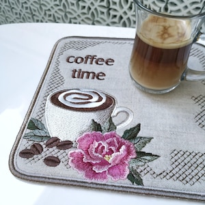 Może przedstawiać: Prostokątna beżowa podkładka z wyhaftowanym napisem coffee time, białą filiżanką kawy z brązowym płynem i pianką, ciemnobrązowymi ziarnami kawy oraz dużym różowym kwiatem z zielonymi liśćmi, na której stoi szklany kubek z kawą.
