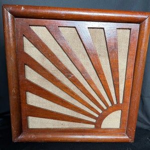 May include: A wooden framed speaker grille with a sunburst design. The grille is made of light brown wood and has a beige fabric backing.