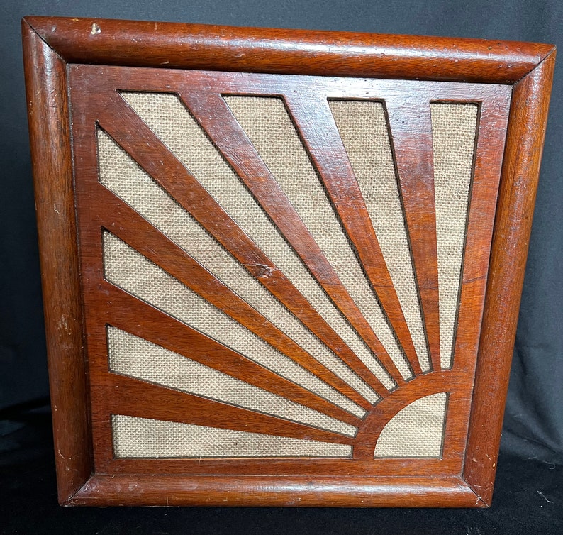 May include: A wooden framed speaker grille with a sunburst design. The grille is made of light brown wood and has a beige fabric backing.