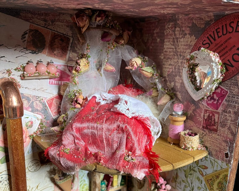 May include: A miniature dollhouse room featuring a red velvet bed with white lace trim and a sheer white curtain. The room is decorated with pink roses, vintage stamps, and a small heart-shaped mirror.