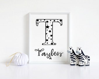 Può includere: Una stampa incorniciata bianca con una lettera nera "T" piena di stelle nere. Il nome "Taylen" è scritto in corsivo nero sotto la lettera. Due figurine di zebra bianche e nere sono in primo piano.